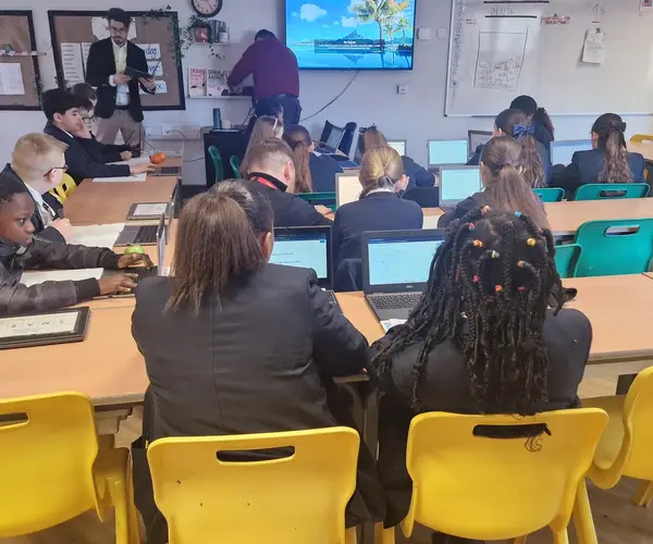 Students in a classroom working on laptops, with a teacher in the background.