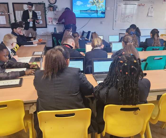 Students in a classroom working on laptops, with a teacher in the background.