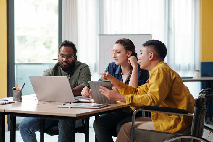 Three individuals engaged in a discussion around a laptop in a bright office setting.