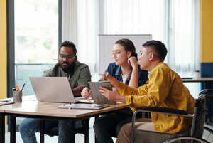 Three individuals engaged in a discussion around a laptop in a bright office setting.