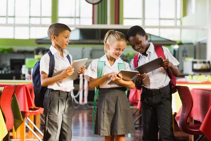 Three schoolchildren are smiling and looking at tablets in a bright cafeteria.