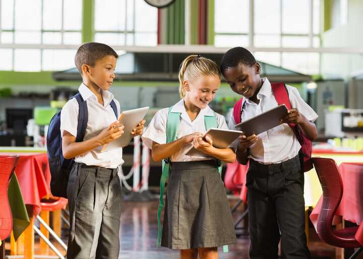 Three schoolchildren with backpacks enjoy using tablets in a bright cafeteria setting.