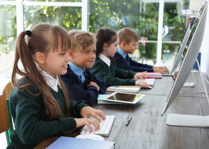 Children sitting at a table, using computers and tablets in a bright classroom.