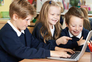 Three children in school uniforms joyfully interact with a laptop in a classroom.