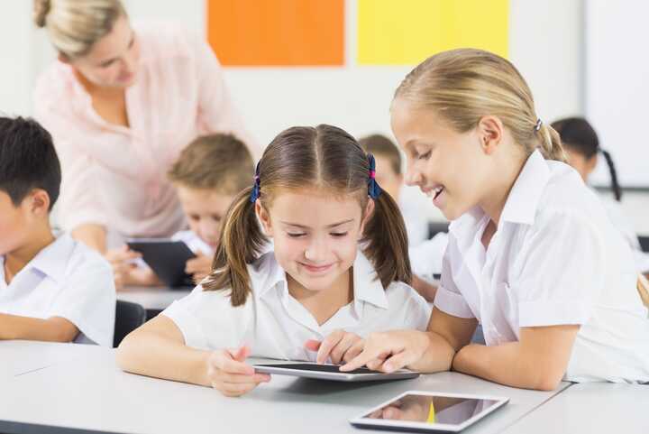 Two girls use a tablet together in a classroom with other students and a teacher in the background.