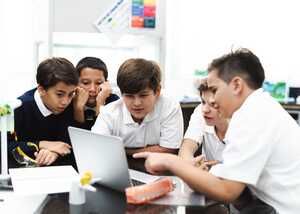 Group of five students leaning over a laptop, engaged in discussion.