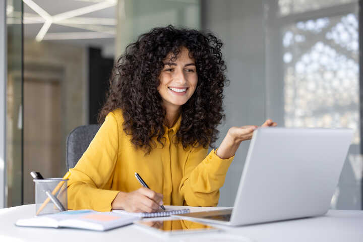 A woman with long hair in a blazer works on a laptop in a classroom setting.