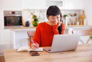 Smiling woman in an orange sweater writing in a notebook while using a laptop.