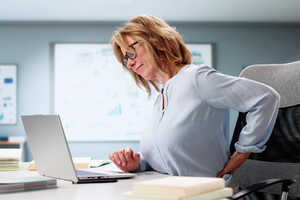 Woman sitting at a desk, looking at a laptop, appears to be in discomfort from back pain.