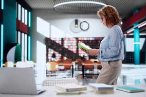 A woman stands at a table with a laptop and books in a modern library setting.