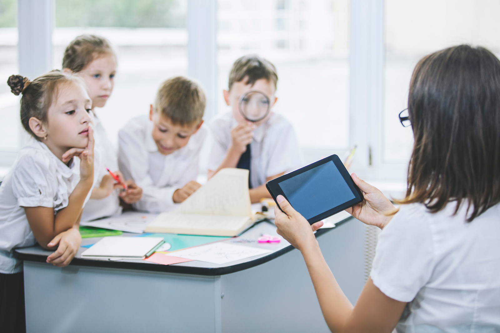A teacher shows a tablet to four attentive children in a classroom setting.