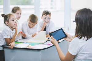 A teacher shows a tablet to four attentive children in a classroom setting.