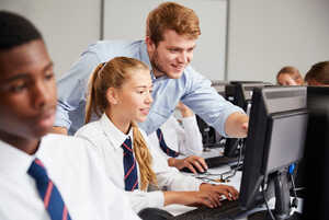 A teacher helps a student at a computer in a classroom setting. Other students are present.
