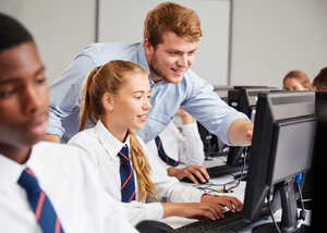 A teacher assisting a student at a computer in a classroom with other students present.
