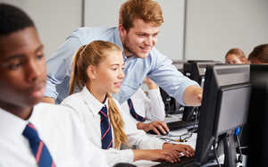 A teacher assists a student at a computer in a classroom with other students present.