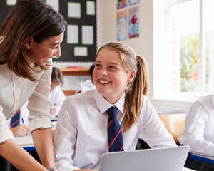 A teacher leans over to assist a smiling student at a laptop in a classroom setting. The student is wearing a white shirt and a tie, while the teacher is dressed in a light-colored blouse. Other students are seen in the background, also engaged