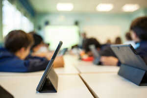 A woman in a blue shirt stands at a table, looking at a laptop in a classroom.