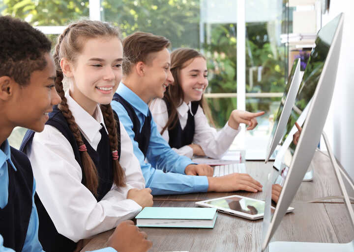 A woman in glasses gestures while sitting at a desk with a laptop and a notebook.