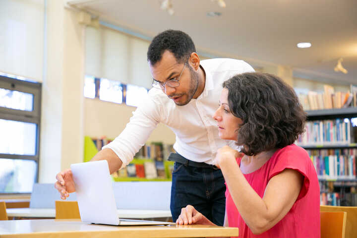 A man helps a woman with a laptop in a library setting.