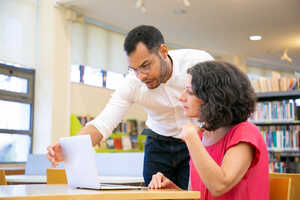 A man helps a woman with a laptop in a library setting.