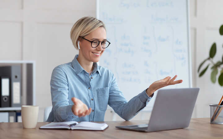 A woman with short blonde hair sits at a desk, gesturing while talking on her laptop.
