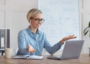 A woman in glasses gestures while sitting at a desk with a laptop and a notebook.