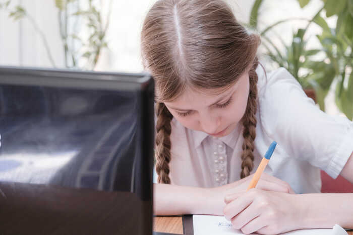 Girl with plaits writing in a school notebook in front of a computer screen