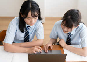 Two students in uniforms focus on a laptop together at a table.