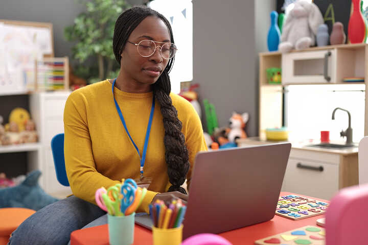 Woman working on a laptop in a colorful classroom setting, surrounded by art supplies.