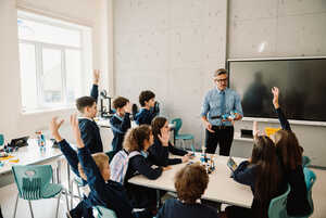 A teacher stands in front of students raising their hands in a classroom setting.