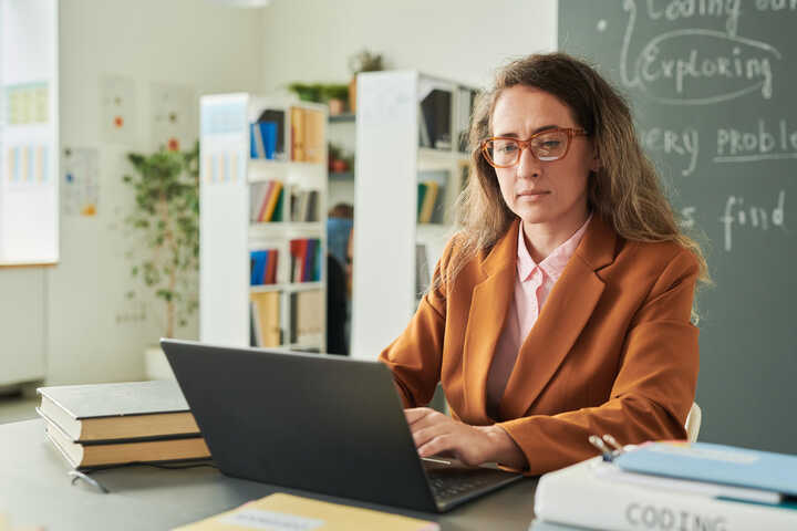 A woman with long hair in a blazer works on a laptop in a classroom setting.
