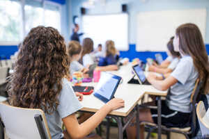 Students in a classroom using tablets, with a teacher presenting in the background.