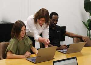 Three people in a meeting room work on laptops, with one pointing at the screen.