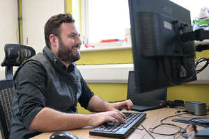 A man with a beard smiles while typing on a keyboard at a desk with monitors.