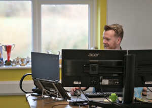 A man smiles while working at a desk with multiple computer monitors in a bright office.