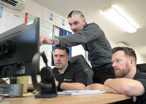 Three men collaborate at a desk, focused on a computer screen in an office setting.