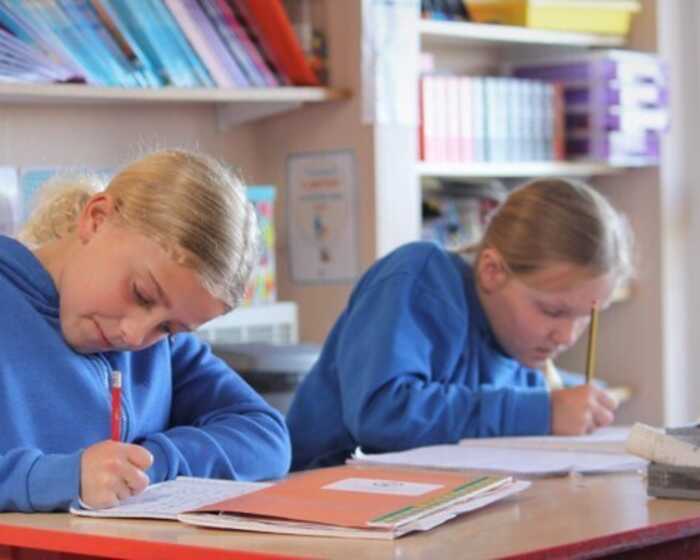 Two children in blue hoodies are focused on writing at a desk in a classroom setting.