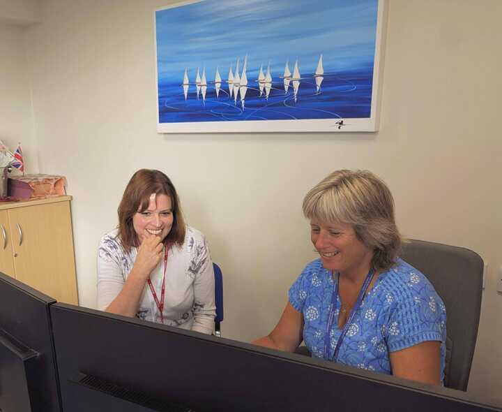 Two women sitting at a desk, looking at a computer monitor with a blue abstract painting above.